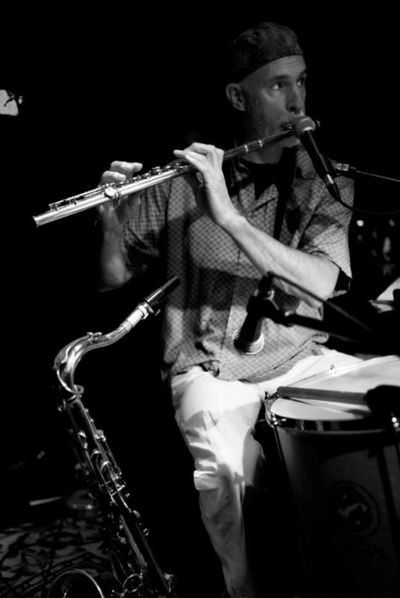 Greg performing with Ginga on a custom flute and Selmer saxophone, seated at a Brazilian surdo drum.