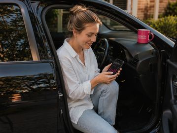 Woman in car doorway checking phone showing car loan details, Coach Brokerage mug on dashboard