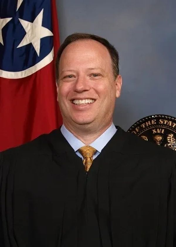 Smiling judge in a black robe with flags in the background.