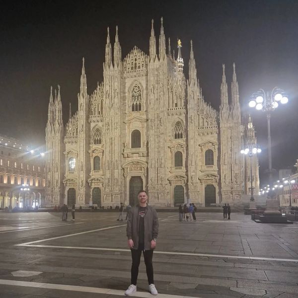 Person standing in front of illuminated Milan Cathedral at night.