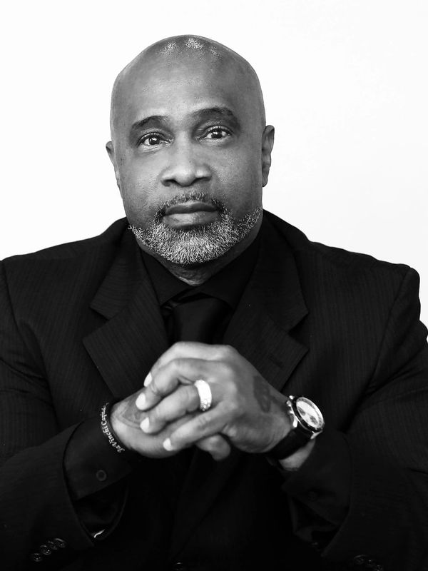 Confident man in a black suit with a contemplative expression, hands clasped.