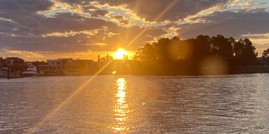 Sunset over a calm river with buildings and trees silhouetted.