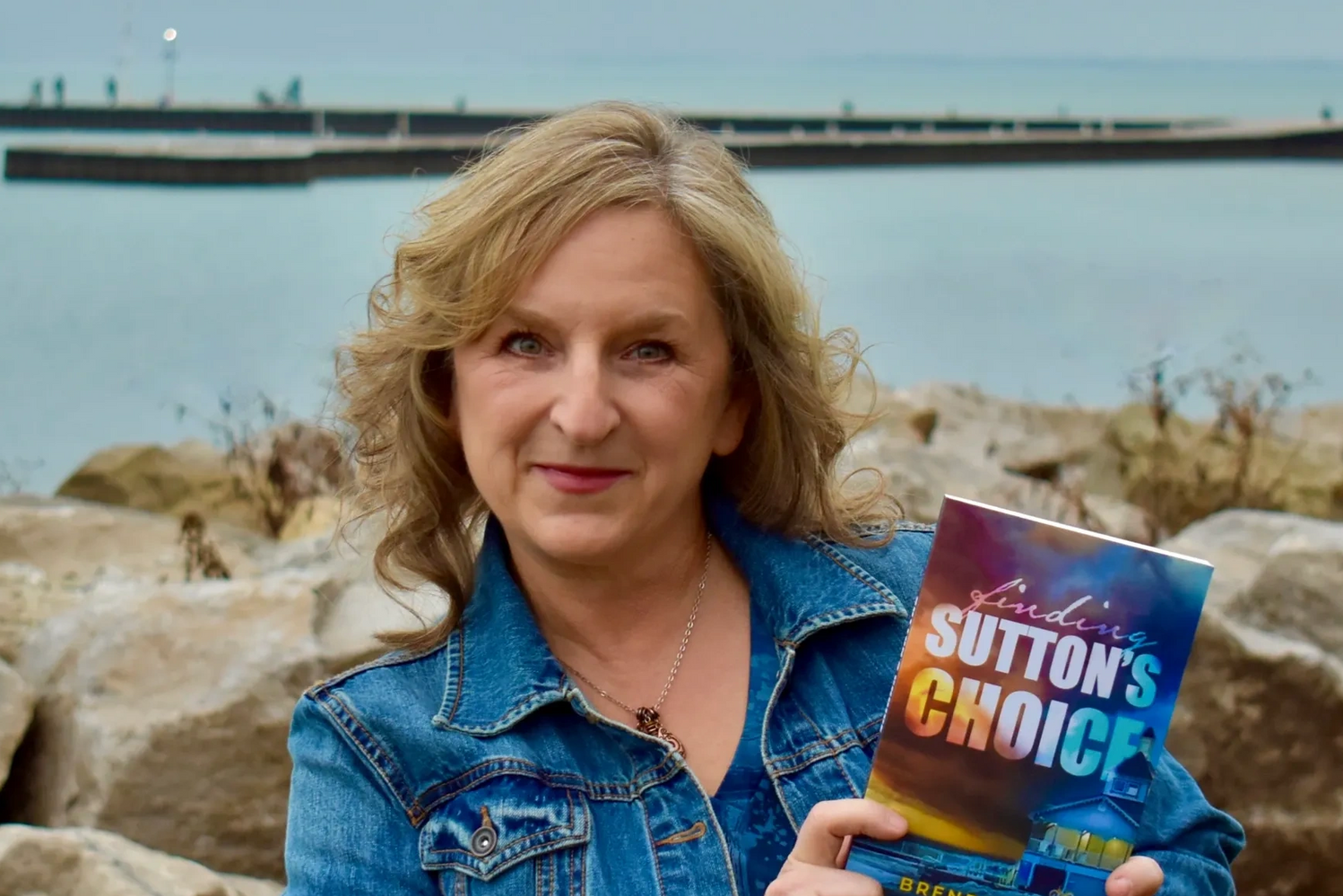 Brenda Haas, a woman holding a book entitled Finding Sutton's Choice, in front of Lake Erie dock