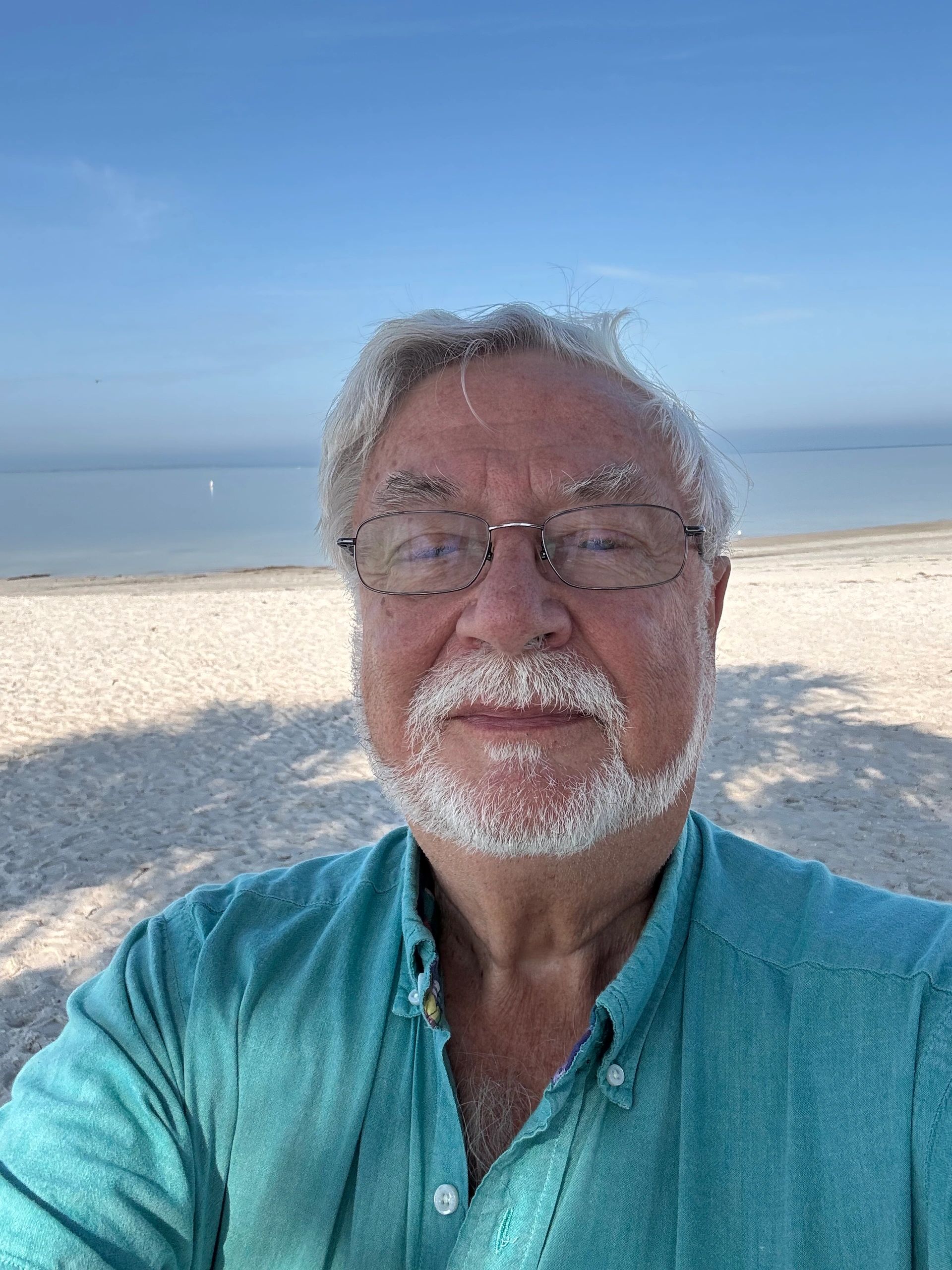 An older man with white hair and glasses takes a selfie on a sunny beach.