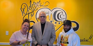 Three people posing in front of a yellow wall with welcoming text and a logo.