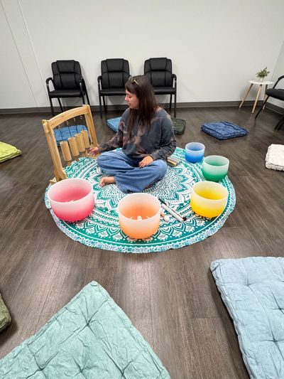Person seated on a patterned mat surrounded by colorful singing bowls.