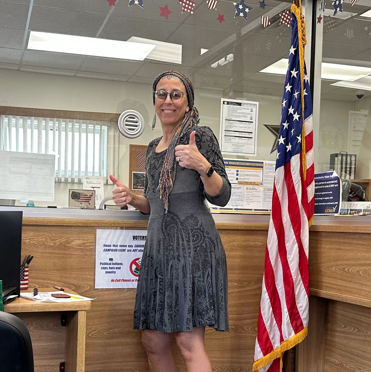 Woman in a gray dress gives thumbs up at a voting location with an American flag nearby.