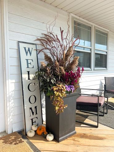 A boho-autumn arrangement with pampas grass. In a black pot, with pumpkins along the bottom. 