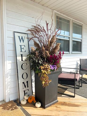 A boho-autumn arrangement with pampas grass. In a black pot, with pumpkins along the bottom. 