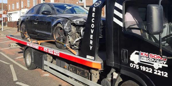 Tow truck carrying a damaged black car on a sunny street.