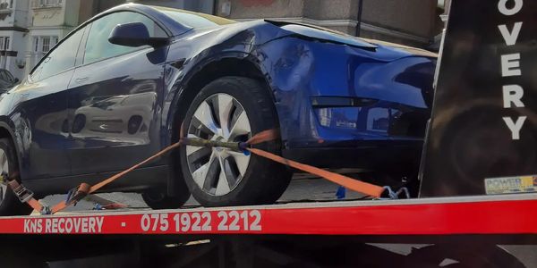 Damaged blue car on a recovery truck in a residential area.
