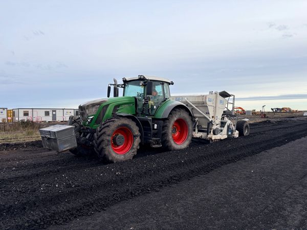 Green tractor with red wheels working on a dark soil or coal surface.