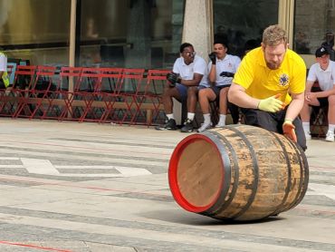 Man rolling a large wooden barrel outdoors.
