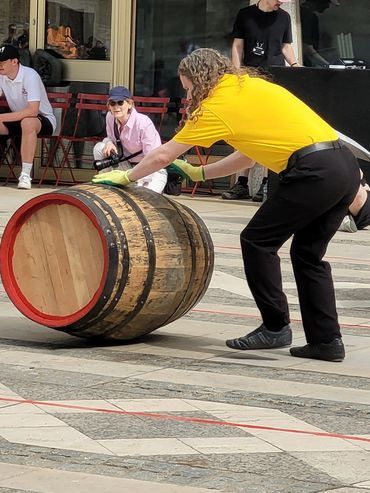Person in yellow shirt rolling a large wooden barrel outdoors.