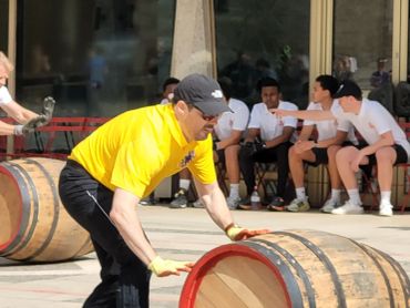 Man in yellow shirt rolling a wine barrel outdoors.