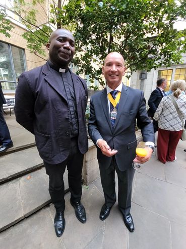 Two men standing outdoors, one in clerical attire and the other in a suit with a medal and a drink.