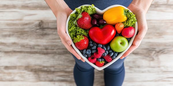 Heart-shaped bowl with fresh fruits and vegetables held by hands.