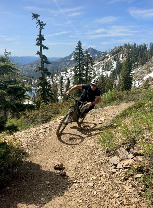 A mountain biker rides on a rocky trail surrounded by trees and mountains.
