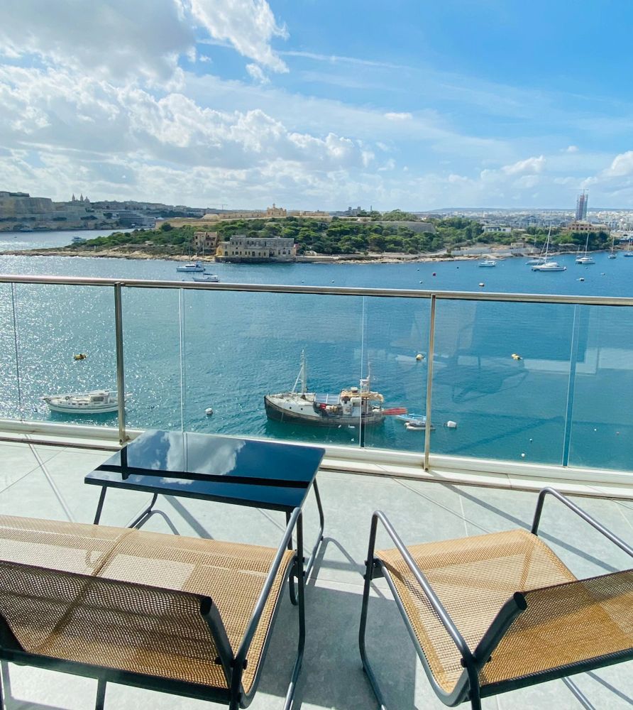 Balcony chairs overlooking a calm blue sea with boats and a sunny sky.