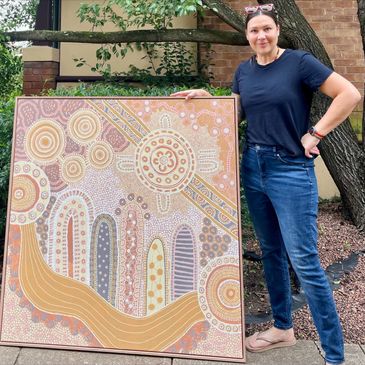 Woman standing beside an Aboriginal-style painting outdoors.