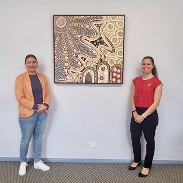 Two women standing beside an Aboriginal art painting on a wall.