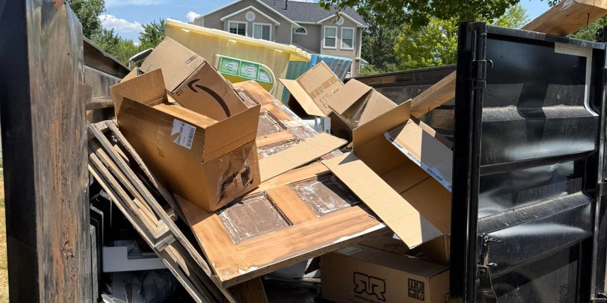 A dumpster filled with cardboard boxes and wooden doors outside a residential house.