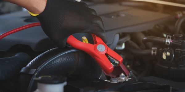 Hand with black glove holding red jumper cable clamp on car battery.