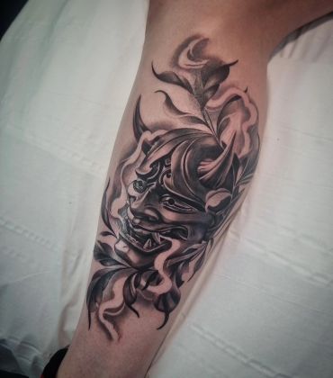 Black and grey tattoo of a demonic mask with swirling smoke and leaves.
