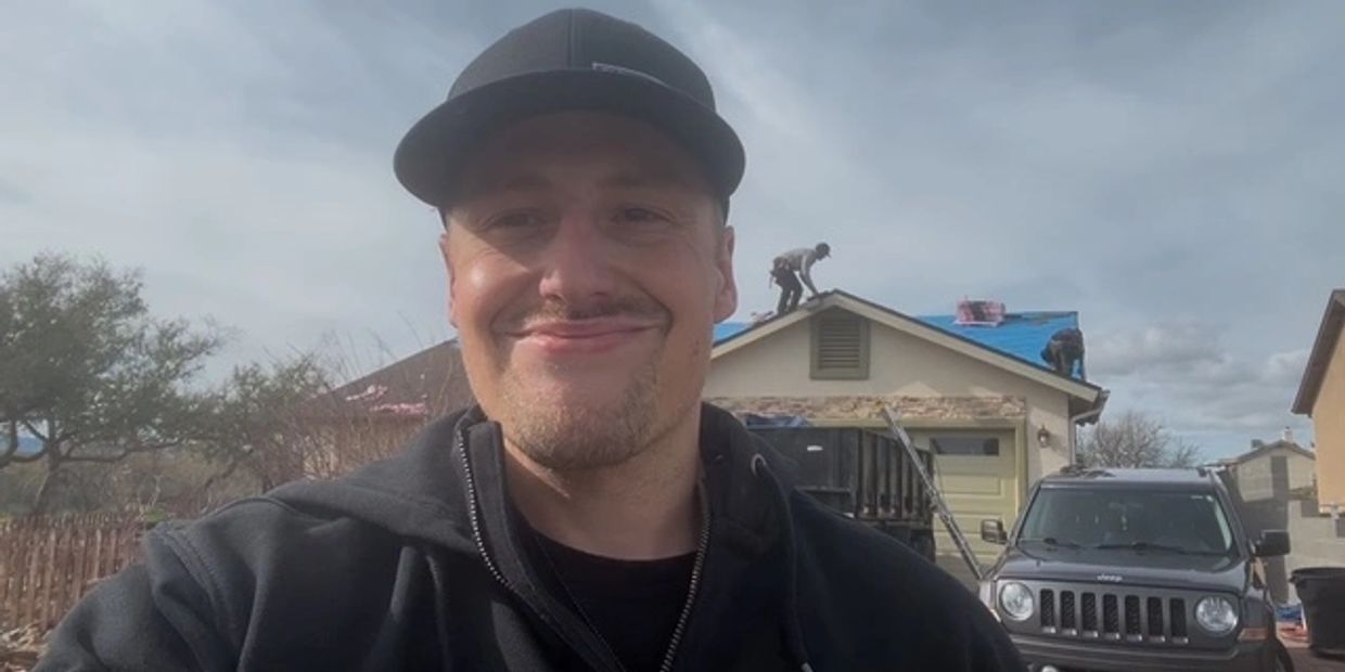 A man in a black cap smiles in front of a house under roof repair with workers on the roof.