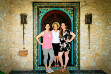Three women standing in front of a colorful tiled archway on a stone wall.