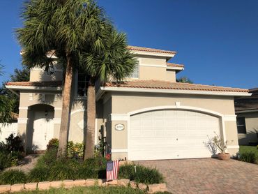 A beige two-story house with a double garage and palm trees in front.