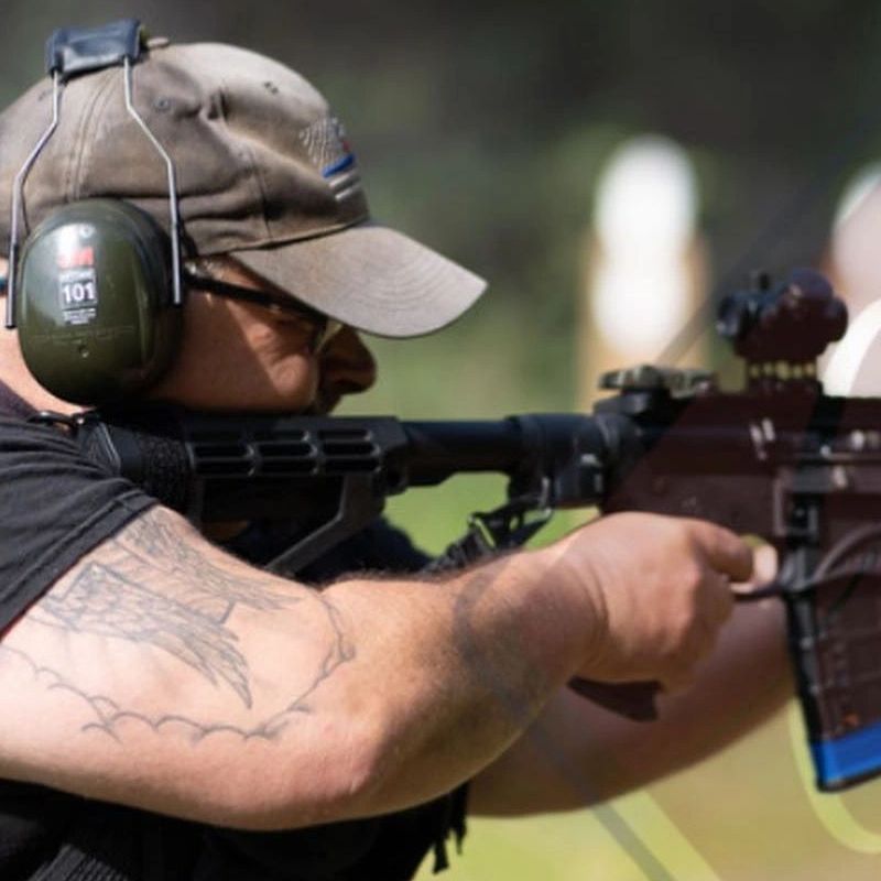 Man wearing hearing protection aiming a rifle at an outdoor shooting range.