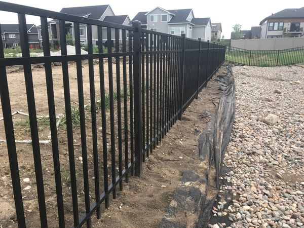 Long black metal fence separating a dirt yard and a gravel area in a suburban neighborhood.