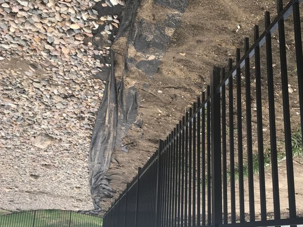 Long black metal fence separating a dirt yard and a gravel area in a suburban neighborhood.