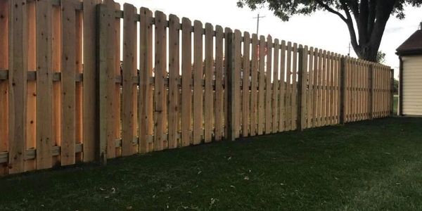 Long wooden fence bordering a grassy yard with a tree and a building.