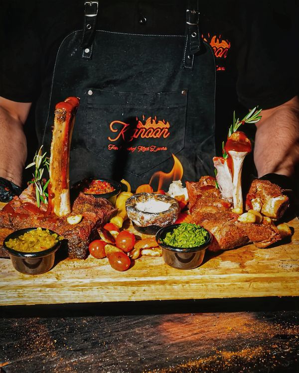 Chef presenting a wooden board with grilled ribs, sauces, and vegetables.