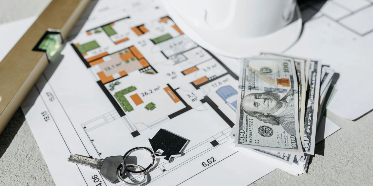 House keys, money, and blueprint with a hard hat on a table.