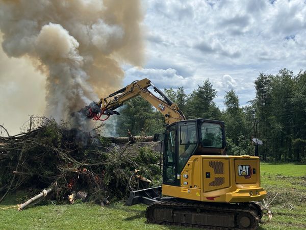 A yellow CAT excavator burning and clearing tree debris in a grassy area.