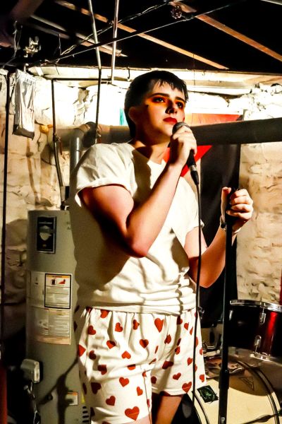 Young person singing into microphone in basement with heart-patterned shorts.