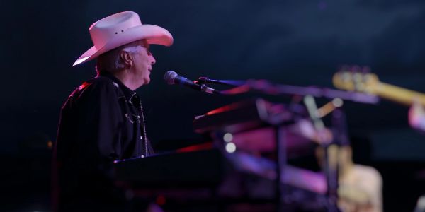 Musician in white cowboy hat singing and playing keyboard on stage.