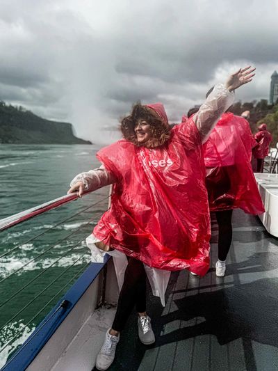 Woman in a red poncho joyfully posing on a boat near a waterfall.