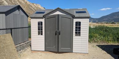 A small gray and white plastic storage shed with double doors and windows in a sunny outdoor area.