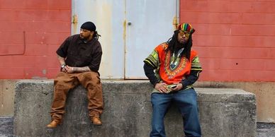 Two men casually sitting against a concrete wall with a red brick background.