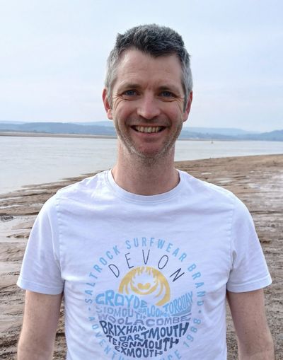 A smiling man in a white Devon-themed t-shirt by the beach.