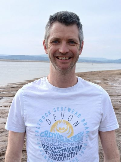 Smiling man in a white graphic t-shirt standing near a beach shore.