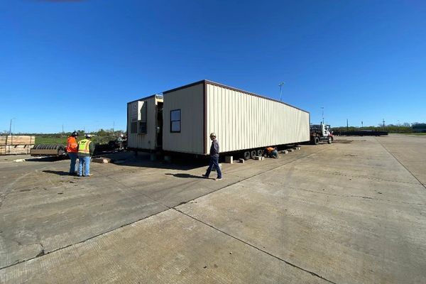Construction workers working around a portable building on a concrete lot under a clear blue sky.