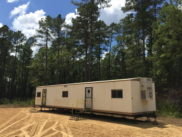 A beige portable building in a forest clearing under a blue sky.