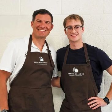 Two men wearing Feeding Broncos aprons smiling together against a white wall.