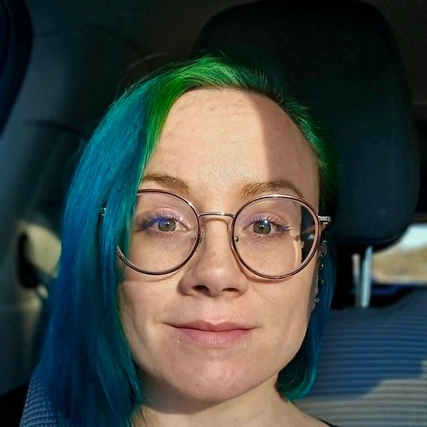 Person with green and blue hair wearing round glasses, sitting in a car.