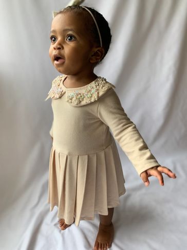 Adorable toddler in a cream dress with beaded collar and headband, standing barefoot.